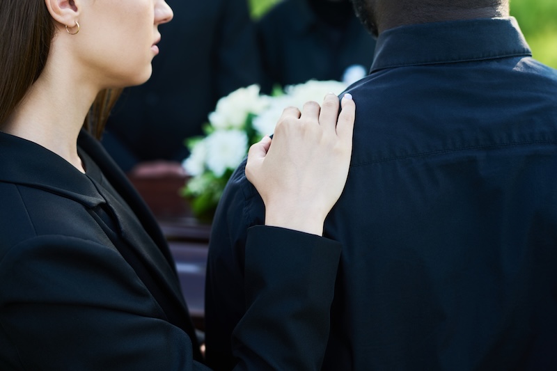 Close-up of young supporting woman consoling her grieving friend wearing mourning clothes at funeral of his wife or other family member