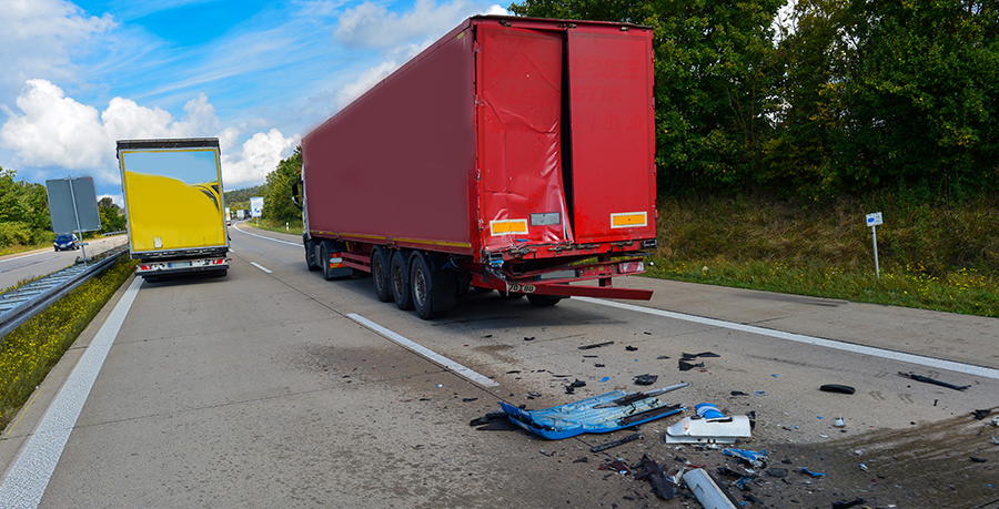 Auffahrunfall auf der Bundesautobahn A7 bei Rothenburg ob der Ta | Law Office of Erik A. Bromson Red cargo trailer damaged at the rear after a crash on a highway, with debris scattered across the road and a second truck in the left lane.